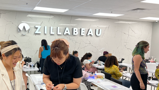 Students gather together with nail art certificates, smiling. A Zillabeau company logo, of a dinosaur, is cast by light is lit on the wall behind them.
